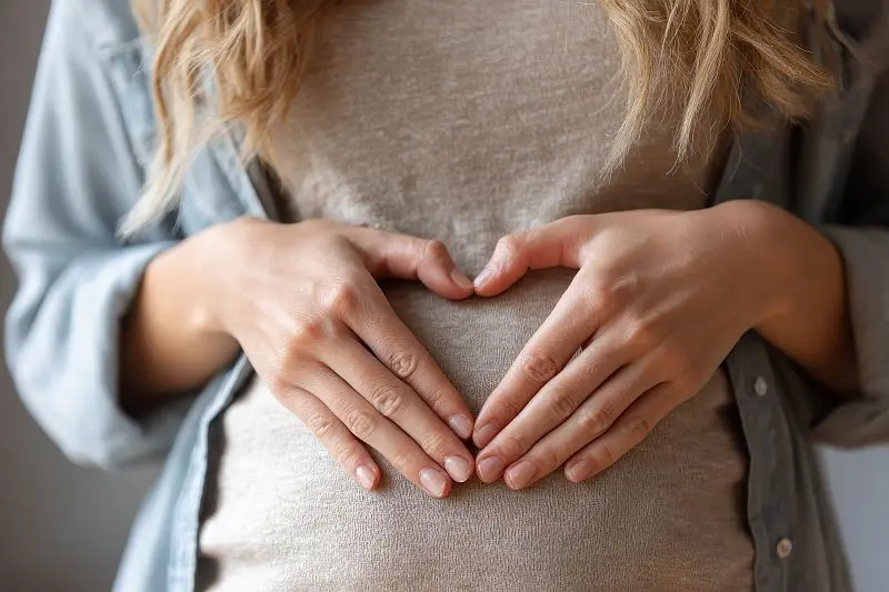 Eine Frau formt ein Herz mit ihren Händen auf ihrem Bauch, symbolisiert durch Frauenheilkunde und spirituelle Verbundenheit.