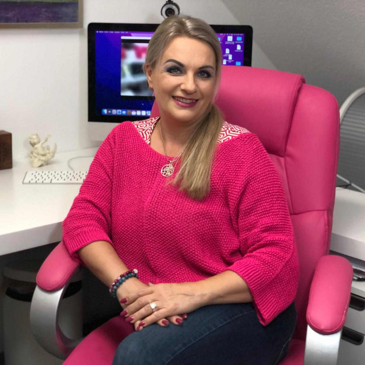 Eine Frau in einem pinken Pullover sitzt lächelnd in einem Büro, bereit für ihre spirituellen Online-Kurse.